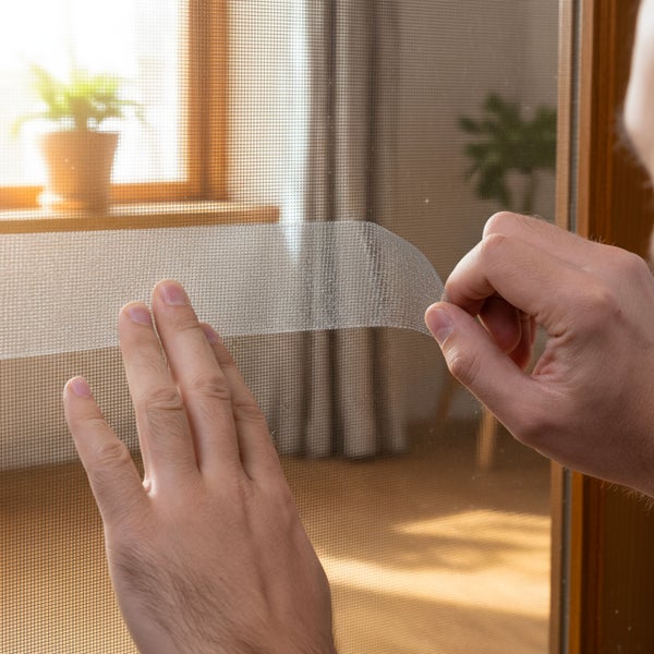 Person befestigt ein Fliegengitter mit Reparaturklebeband an einem Fensterrahmen.