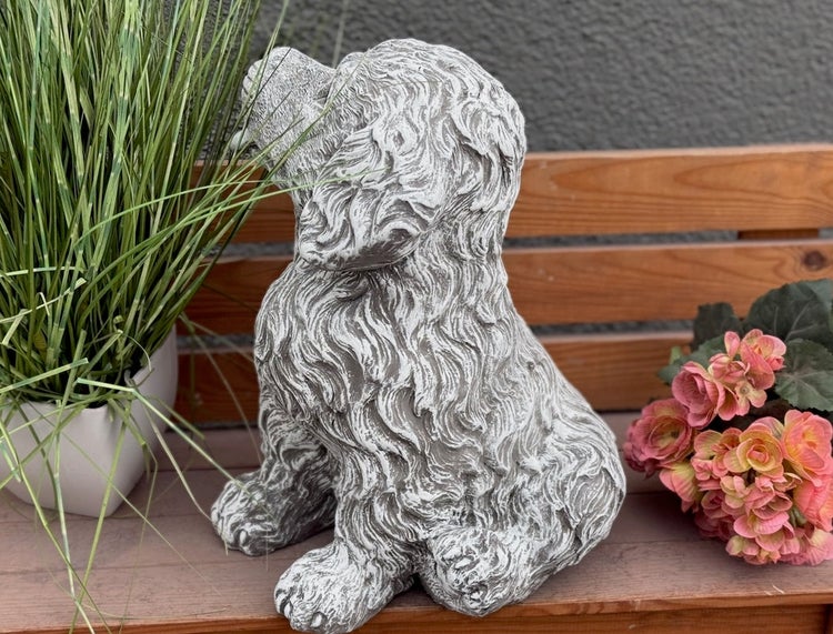 Gartenfigur in Form eines sitzenden Hundes aus Steinguss, auf einer Holzbank platziert neben Gras und Blumen.