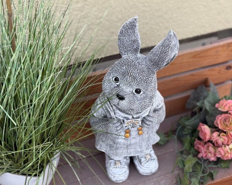 Gartenfigur Hase mit Kleidung und Schuhen, stehend auf einer Holzbank neben einer Pflanze.