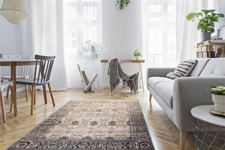 Helles Wohnzimmer mit einem gemusterten Orientteppich auf Fischgrätparkett, grauem Sofa, Esstisch und Schreibtisch vor hellen Fenstern.