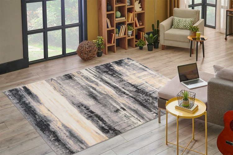 Teppich mit abstraktem Muster in Grau, Schwarz und Creme auf hellem Holzboden in einem hellen Wohnzimmer mit Bücherregal und Sessel.