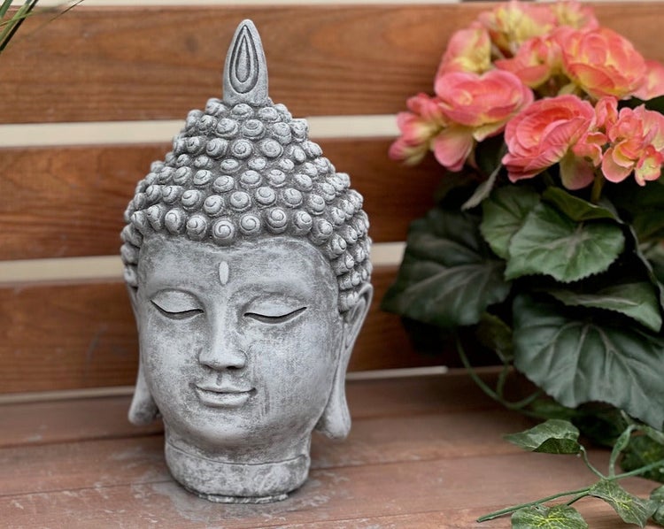Buddha-Kopf Gartenfigur in Steinoptik auf einer Holzfläche neben einer Pflanze.