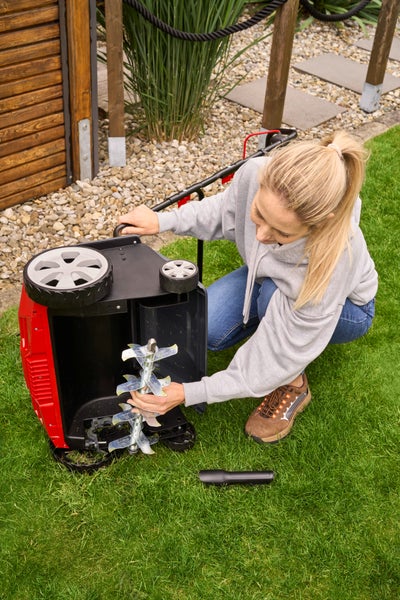 Eine Frau kniet auf einer Rasenfläche und wechselt die Vertikutierwalze an einem roten Elektro-Vertikutierer in einer hellen Gartenanlage.