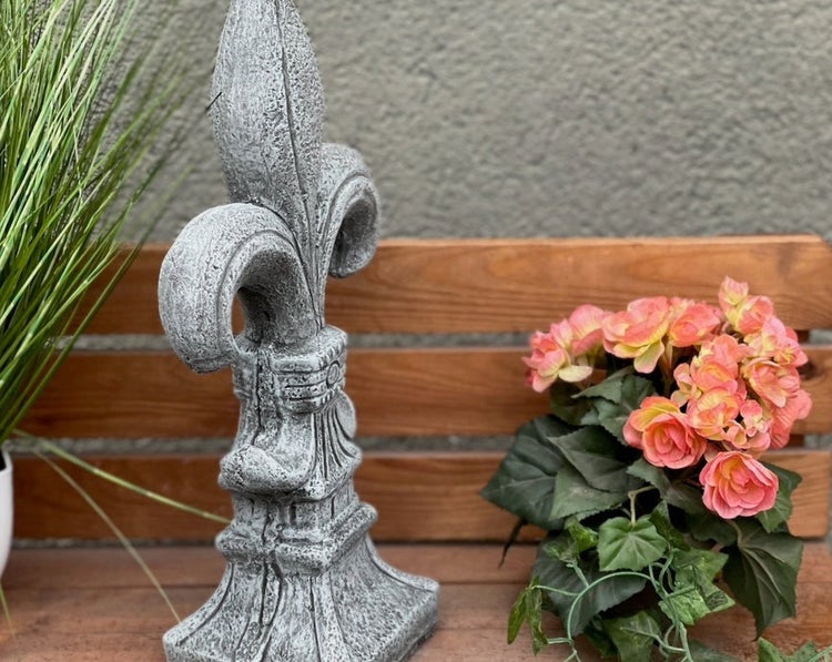 Gartenfigur französische Lilie in Steinoptik auf einer Holzbank neben Blumen.