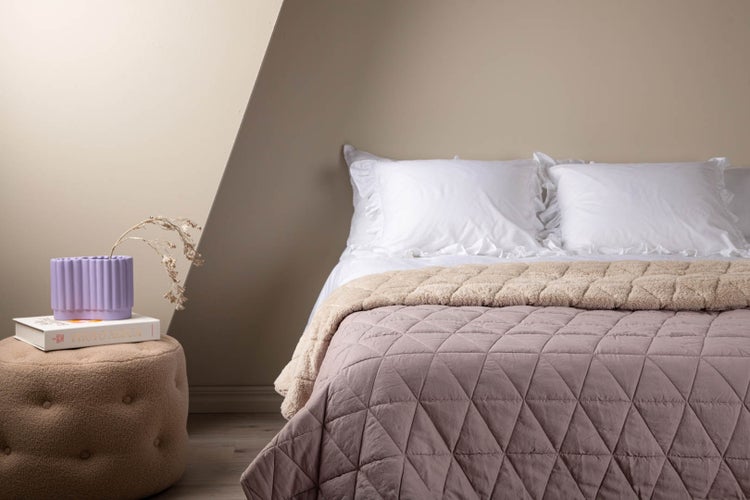Gemütliches Schlafzimmer mit malvenfarbener gesteppter Tagesdecke mit Dreiecksmuster, weißen Rüschenkissen und braunem Teddy-Puff.