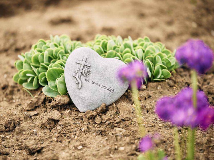 Herzfoermiger Grabstein mit Kreuz und Rose sowie der Inschrift Wir vermissen dich als Grabschmuck im Garten