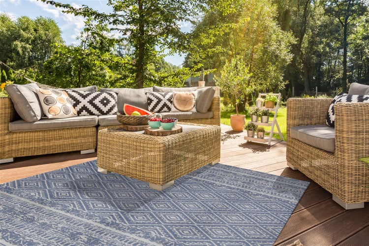 Helle Holzterrasse mit Polyrattan-Gartenmöbeln und blauem Outdoorteppich mit geometrischem Muster, umgeben von Bäumen im Garten.