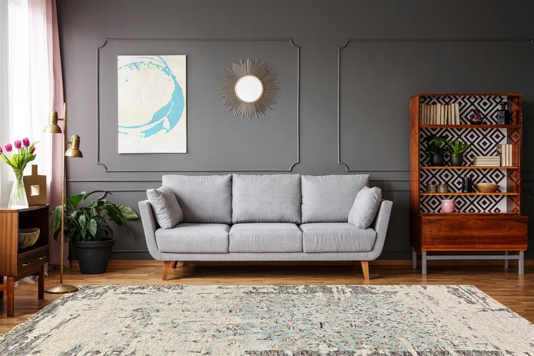 Wohnzimmer mit grauem Dreisitzer-Sofa, gemustertem Teppich, Holzregal und dunkelgrauer Wand mit Zierleisten.