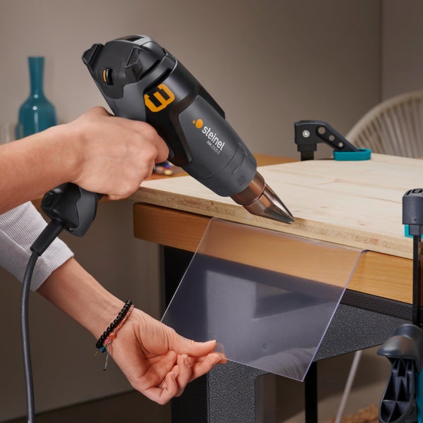 Eine Hand hält eine Steinel Heißluftgebläse HM 2020 E, während die andere Hand ein Kunststoffblatt an einer Holzkante bearbeitet.