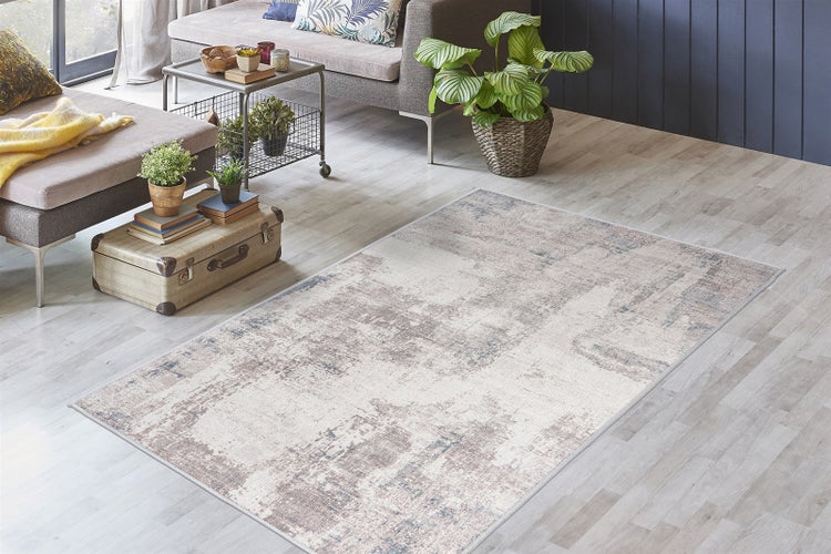 Rechteckiger Teppich mit abstraktem Muster in einem hellen Wohnzimmer auf hellem Laminatboden neben grauen Sofas und Zimmerpflanzen.