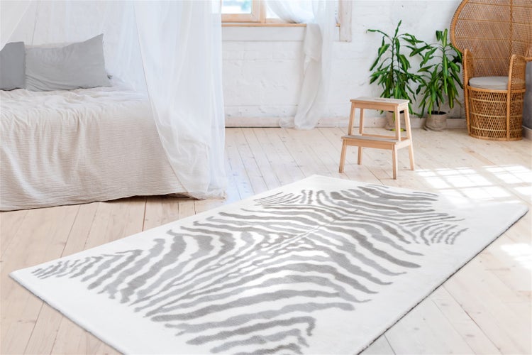 Rechteckiger Teppich mit grau-weißem Zebramuster auf hellem Holzboden in einem hellen Zimmer mit Bett und Pflanzen.