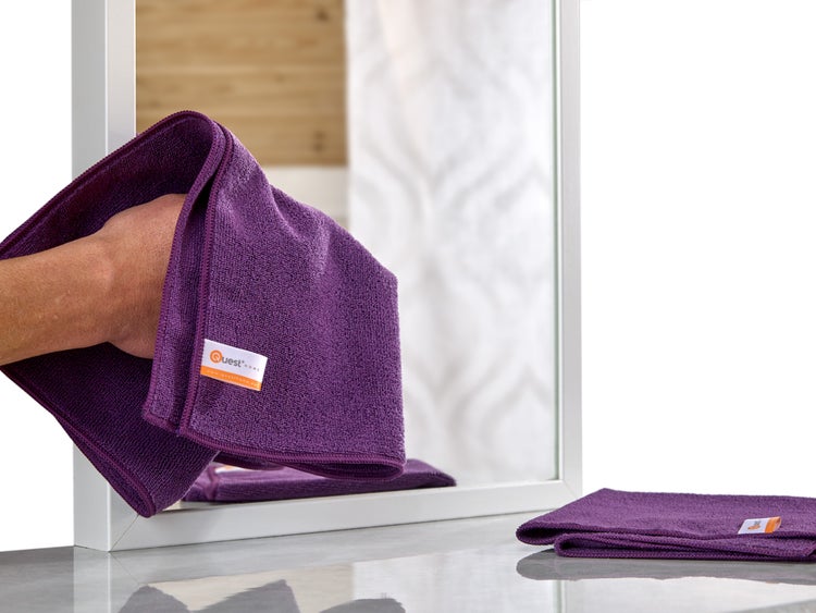 Eine Person reinigt einen Spiegel mit einem violetten Mikrofasertuch; ein weiteres Tuch liegt auf der Oberfläche