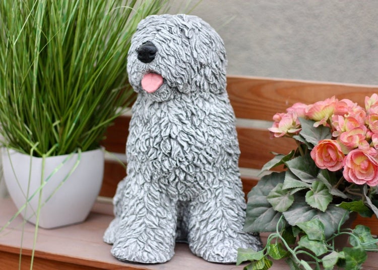 Gartenfigur in Form eines sitzenden, zotteligen Hundes aus Kunststein auf einer Holzbank zwischen Pflanzen.