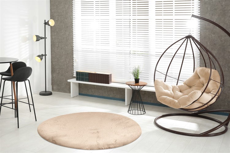 Helles modernes Wohnzimmer mit rundem beigem Teppich, dunklem Hängesessel, Fenster mit weißen Jalousien und dreiflammiger Stehleuchte.