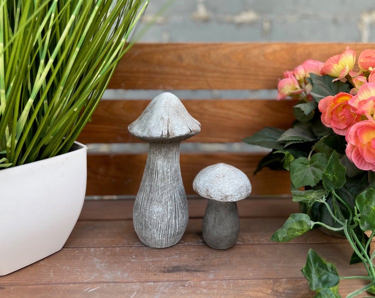 Zwei Deko-Pilze in Steinoptik auf einer Holzbank, umgeben von Pflanzen und Blumen.