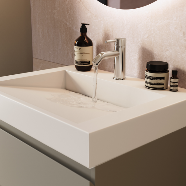 Modernes Waschbecken mit Armatur und fließendem Wasser, ergänzt durch Pflegeprodukte auf der Ablagefläche vor einem beleuchteten Spiegel.