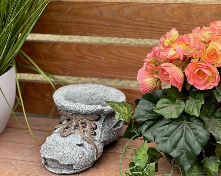 Pflanztopf in Form eines Schuhs aus Steinguss für die Gartendekoration, auf einer Holzoberfläche neben einer Pflanze.