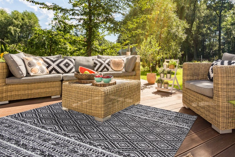 Garten-Lounge aus Polyrattan mit Ecksofa, Sessel, Tisch, grauen Polstern und geometrischem Outdoor-Teppich auf Holzterrasse im sonnigen Garten.