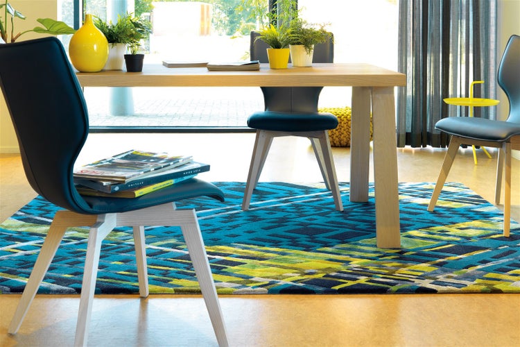 Hell gestaltetes Zimmer mit Holztisch, blauen Polsterstühlen und einem Teppich mit geometrischem Muster in Blau, Grün und Gelb.