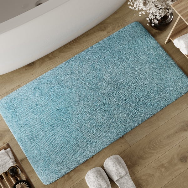 Rechteckige blaue Badematte aus weichem Material auf Holzboden neben einer Badewanne mit Hausschuhen und Handtüchern.