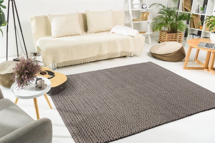 Rechteckiger dunkelgrauer Webteppich in einem hellen, modern eingerichteten Wohnzimmer mit hellem Sofa, Holzmöbeln und Zimmerpflanzen.