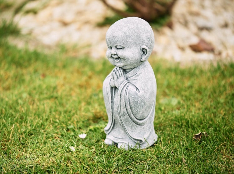 Steinfigur kleiner lachender Buddha im Gebet auf einer Wiese stehend für die Gartendekoration