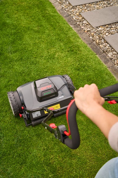 Akku-Spindelmäher von AL-KO auf einer hellen, gepflegten Rasenfläche neben einem Steinweg, ergonomisch geformter Griff wird von einer Hand geführt.