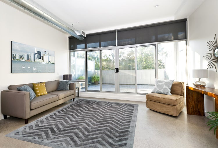 Helles Wohnzimmer mit großer Glas-Schiebetür, dunklen Rollos, grauem Sofa, Teppich mit Zickzack-Muster und sichtbarem Lüftungsrohr im Loft-Stil.