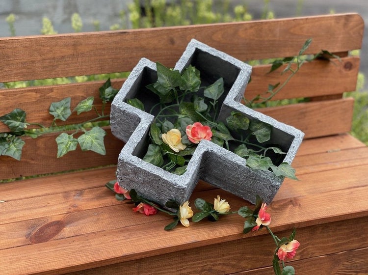Grabschale in Kreuzform aus steinerner Optik dekoriert mit künstlichen Efeu und Blumen auf einer Holzbank.