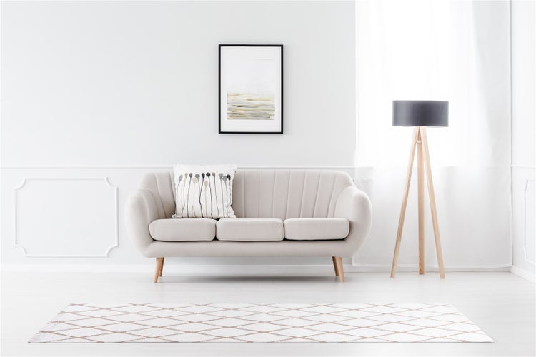 Helles Wohnzimmer mit beigem Sofa, Dreibein-Stehlampe aus Holz, gemustertem Teppich und abstraktem Wandbild in minimalistischem Stil.