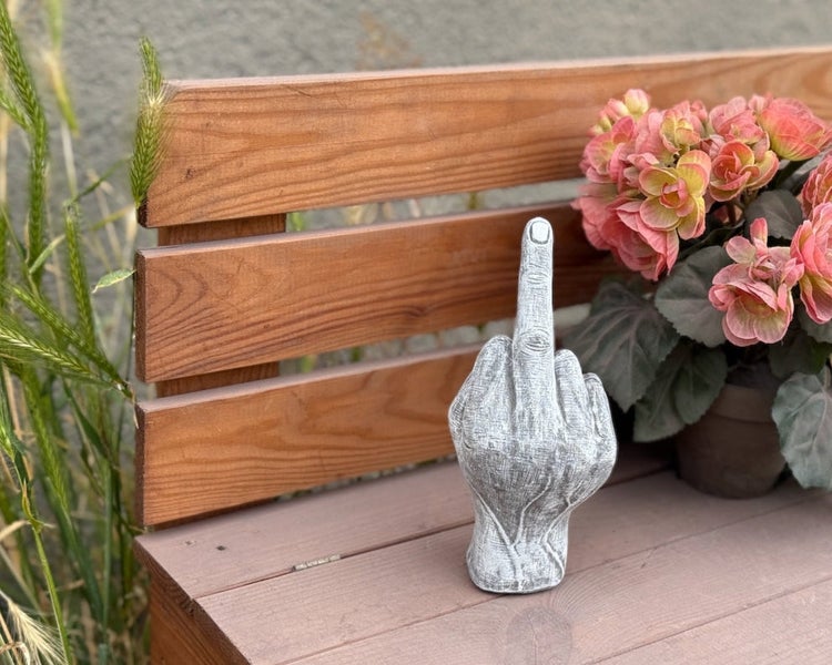 Dekorative Gartenfigur aus Stein in Form einer erhobenen Hand mit ausgestrecktem Mittelfinger, platziert auf einer Holzbank neben Blumen.