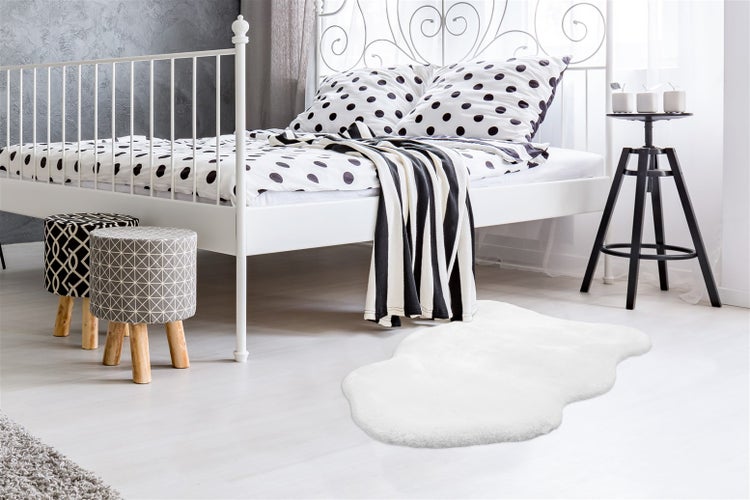Helles Schlafzimmer mit weißem Metallbett, gepunkteter Bettwäsche, weißem Kunstfellteppich, zwei gemusterten Hockern und schwarzem Beistelltisch.