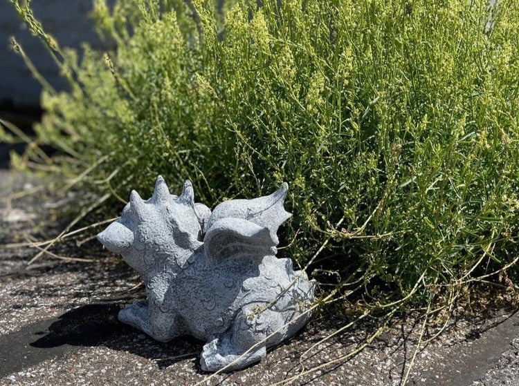 Dekorative Gartenfigur in Drachenform aus Stein, platziert in einem Gartenbeet vor einer Pflanze.