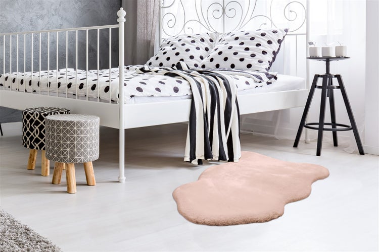 Hell gestaltetes Schlafzimmer mit weißem Metallbett, Bettwäsche mit Punktemuster, zwei gemusterten Hockern und einem rosa Kunstfellteppich.