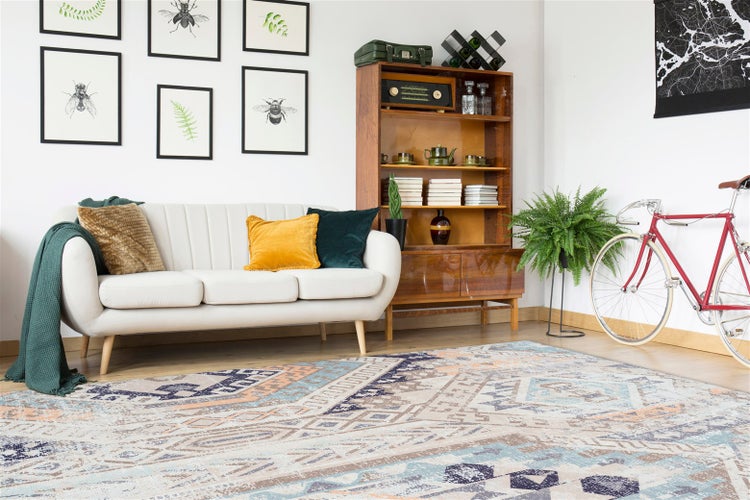 Hell gestaltetes Wohnzimmer mit weißem Sofa, buntem Teppich mit geometrischem Muster, Holzregal, Wandbildern und einem roten Fahrrad.