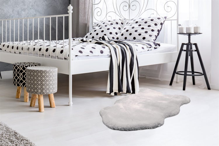 Hellgrauer Kunstfellteppich in hellem, modernem Schlafzimmer mit weißem Metallbett, gepunkteter Bettwäsche und gemusterten Holzhockern.