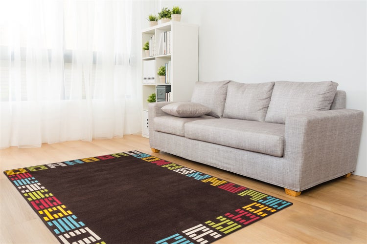 Modern eingerichtetes Wohnzimmer mit grauem Stoffsofa, weißem Standregal und einem dunkelbraunen Teppich mit buntem geometrischem Muster auf Laminat.