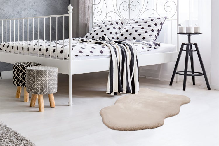 Schlafzimmer mit weißem Metallbett, gepunkteter Bettwäsche, zwei Hockern und einem beigen Kunstfellteppich auf hellem Holzboden.
