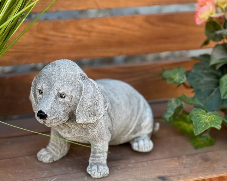 Gartenfigur in Form eines Dackels aus Steinguss, sitzend auf einer Holzbank.