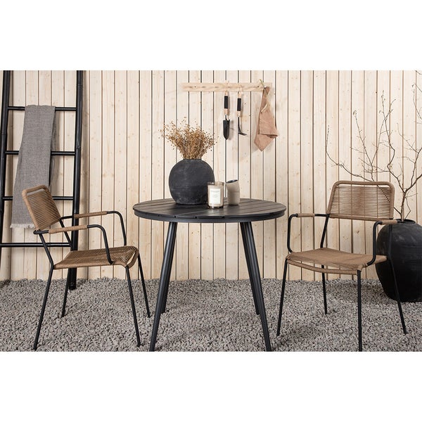 Gartenszene mit rundem Tisch, zwei Stühlen, Deko-Objekten und Gartengeräten vor einer Holzwand