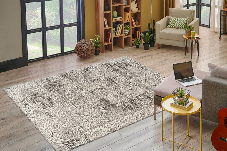 Großer Teppich mit Vintage-Muster in Grau und Weiß auf Holzboden in hellem Wohnzimmer mit Bücherregal, Sessel und großem Fenster.