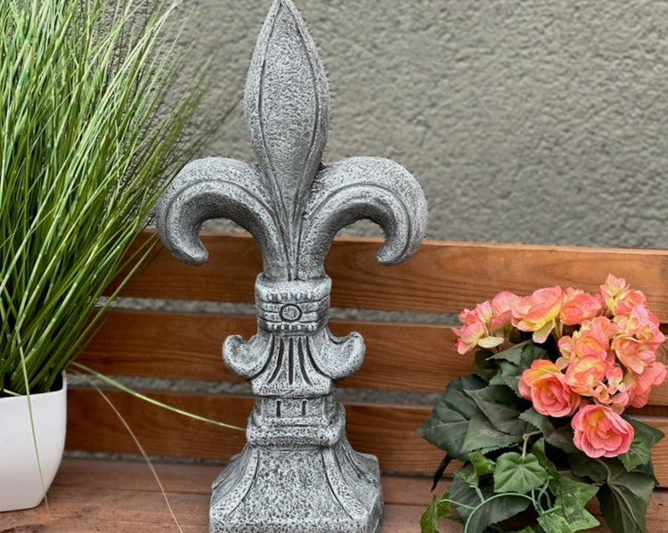 Gartenfigur Fleur-de-Lis aus Steinguss auf einer Holzbank neben Topfpflanzen.
