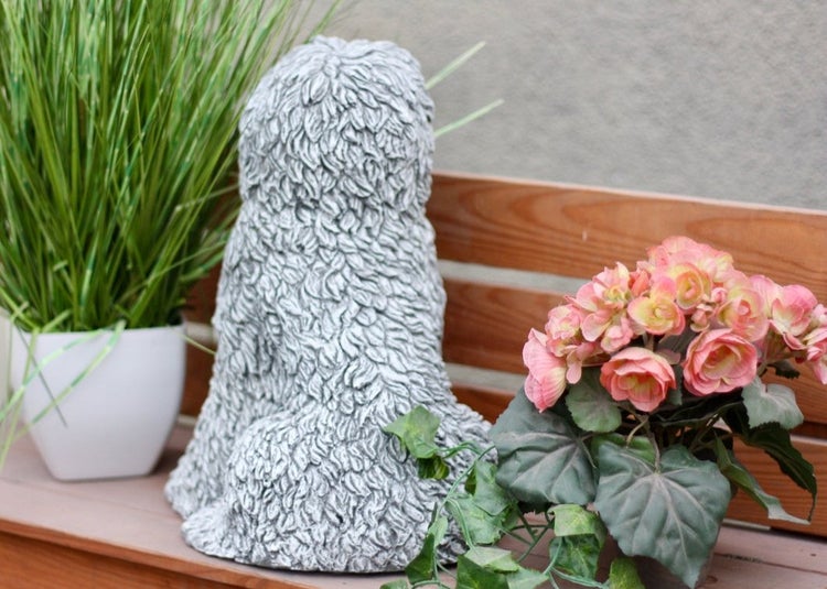 Gartenfigur aus Stein in Form eines sitzenden Pudels von hinten, platziert auf einer Holzbank neben Topfpflanzen.