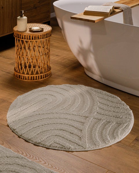 Runder strukturierter Badevorleger mit Wellenmuster in Bad mit freistehender Badewanne, Holzboden und dekorativem Beistelltisch aus Rattan.