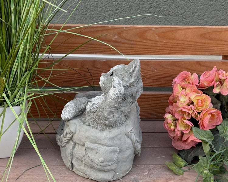 Gartenfigur einer Katze in einer Tasche in Steinoptik, platziert auf einer Holzbank neben Pflanzen und Blumen.