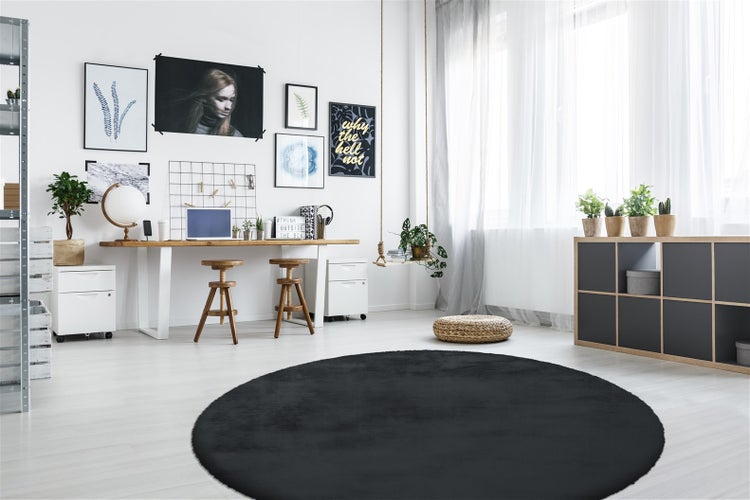 Runder schwarzer Teppich in einem hellen, modern eingerichteten Arbeitszimmer mit Holzschreibtisch, Hockern und Wandbildern.