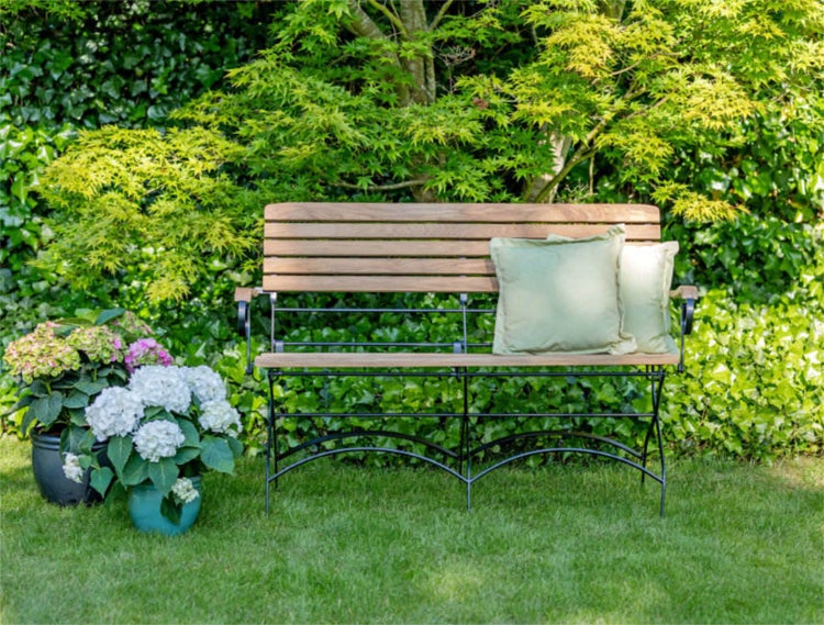 Gartenbank aus Holz und Metall mit Kissen vor grüner Gartenkulisse