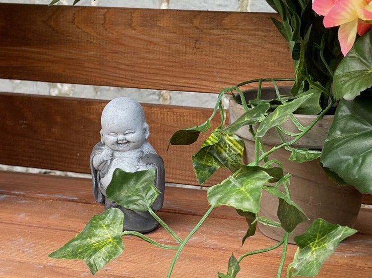 Kleine lachende Buddha Figur aus Stein in einer dekorativen Gartenumgebung auf einer Holzbank mit Pflanze.