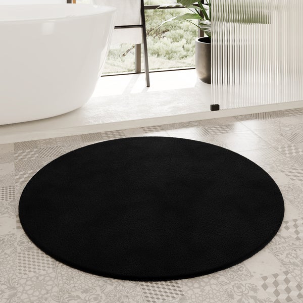 Runder schwarzer Badeteppich auf gemustertem Fliesenboden in einem hellen Badezimmer mit freistehender Badewanne und Glasduschwand.