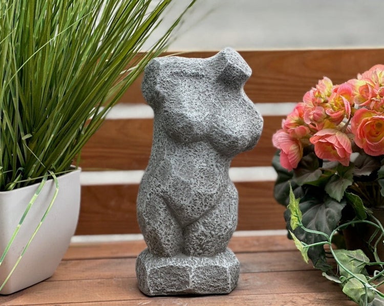 Abstrakte Gartenfigur in Form eines weiblichen Torsos mit rauer Steinoptik, platziert auf einem Holztisch neben Pflanzen.